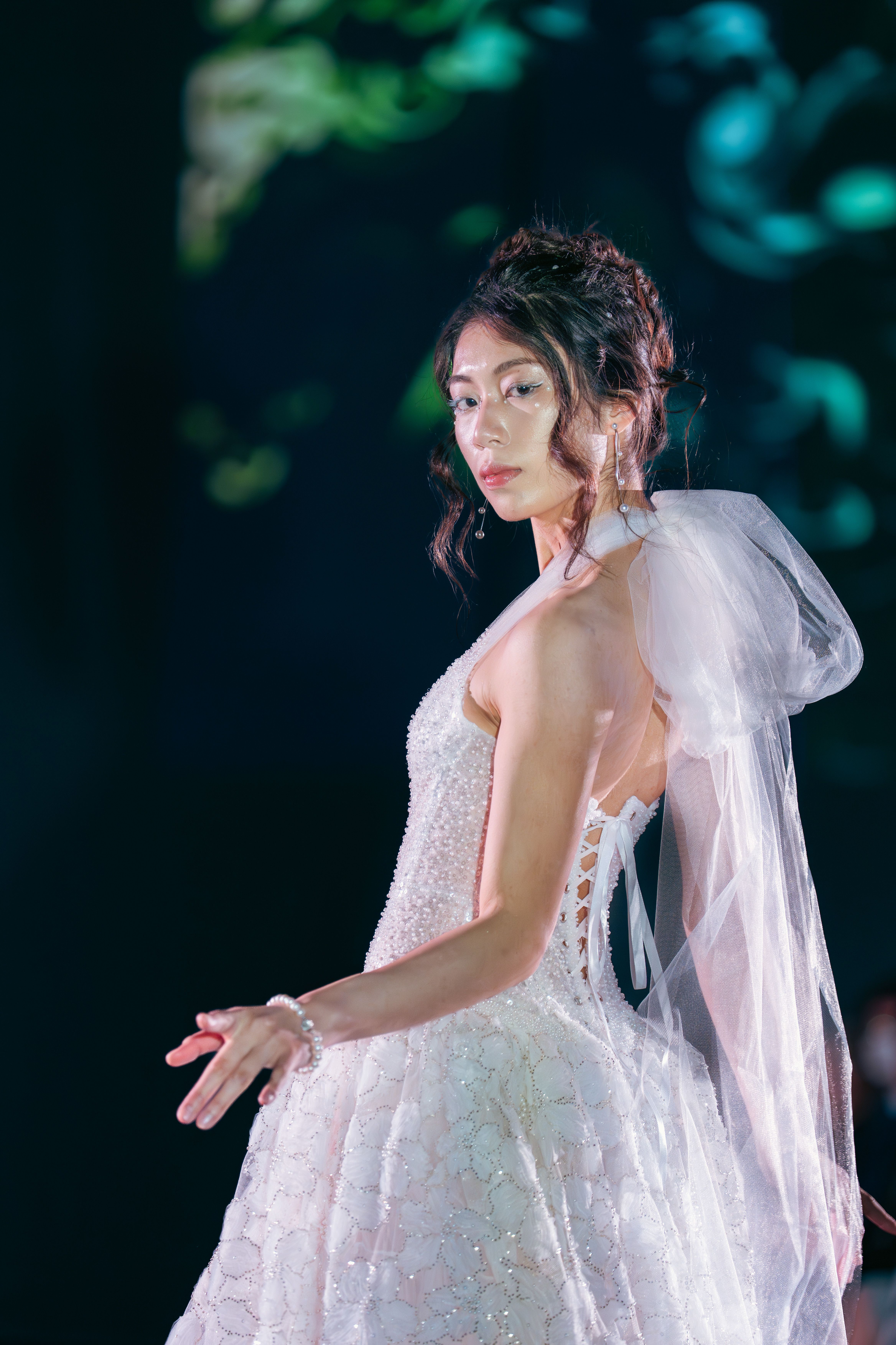 Model wears a sleeveless white gown adorned with pearls and floral embroidery. The dress laces up at the side and features a large sheer bow at the shoulder. She accessorizes with pearl jewelry.